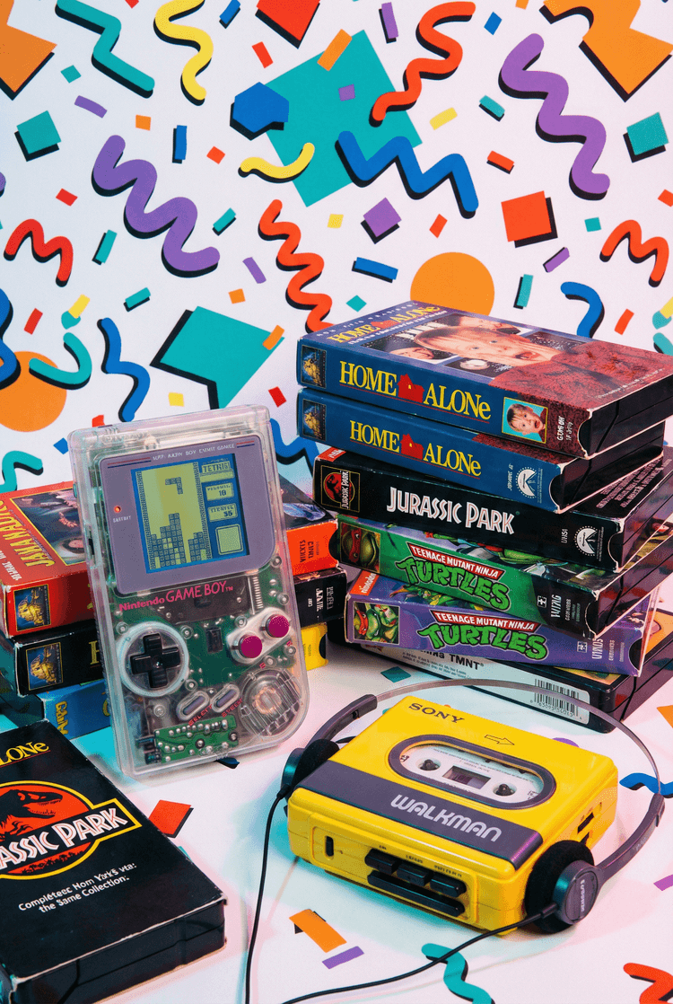 A still life of 1990s nostalgia items including a clear plastic Gameboy, a pile of VHS tapes, and a walkman, vibrant mem
