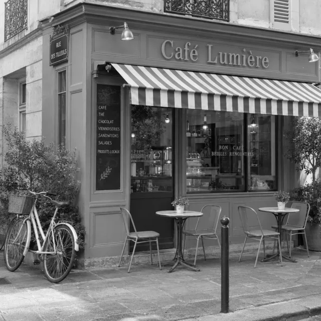 Black and white photo of a corner cafe with a striped awning, a sign reading Café Lumière, chairs and a bicycle