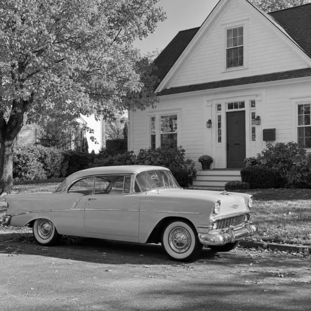 Black and white photo of a 1950s car parked in front of a white house with a tree