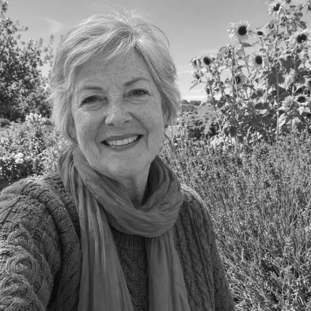 Black and white photo of a fictional woman with silver hair in a sweater and scarf in a garden with sunflowers