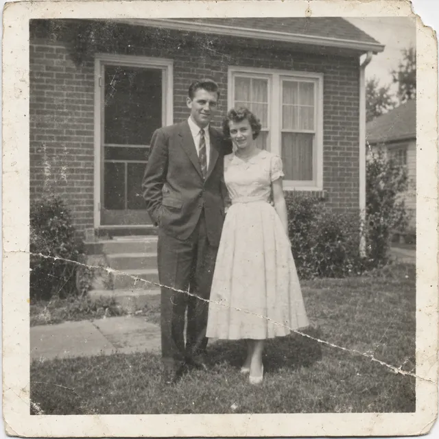 Original photo: faded black-and-white print of a couple in front of a brick house, with scratches, a diagonal crease and a torn corner