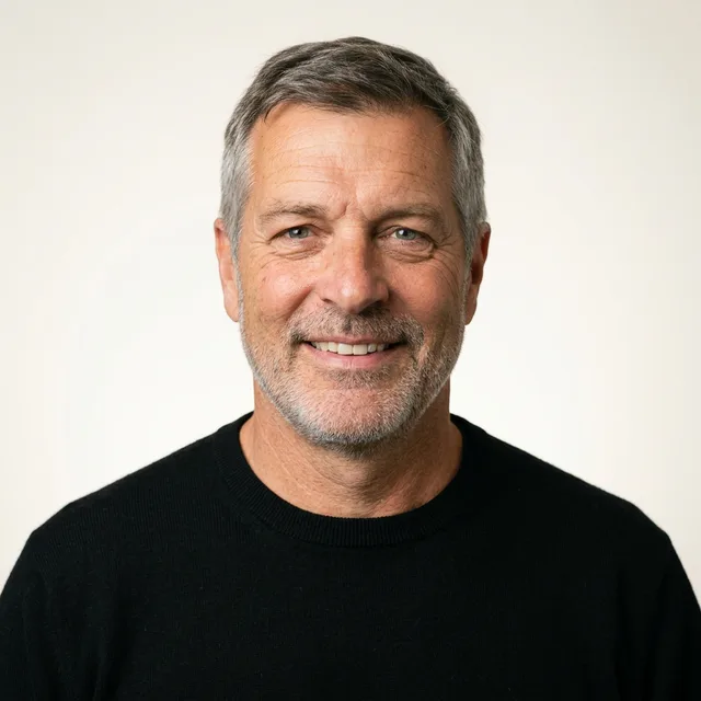 The grey-haired man in a black crew-neck sweater on the same warm white backdrop