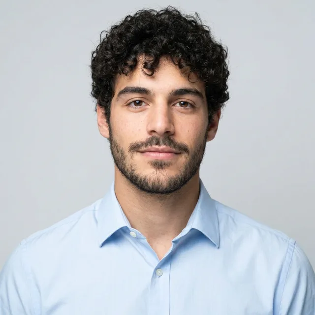 The same young man with curly hair in a light blue shirt on light grey, with a thinner, neater beard
