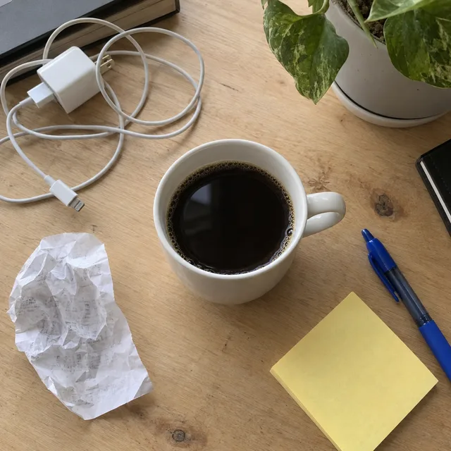 Synthetic source photo: a mug of coffee on a wooden desk with a charger, cable, crumpled receipt, sticky note, pen and plant