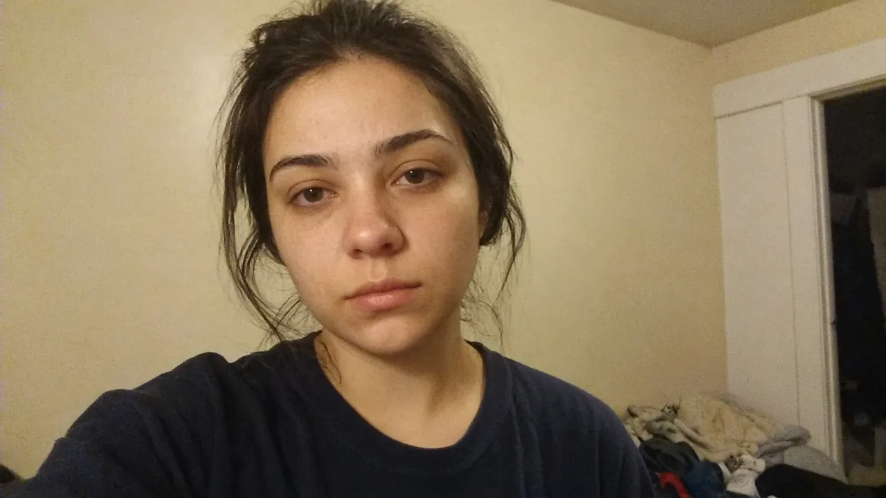 Indoor selfie of a woman with a tired expression and dark circles against a plain wall